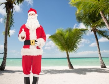 christmas, holidays, gesture and people concept- man in costume of santa claus showing thumbs up over tropical beach background