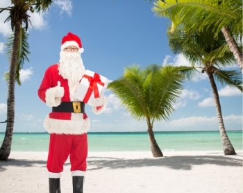 christmas, holidays and people concept - man in costume of santa claus with gift box showing thumbs up gesture over tropical beach background