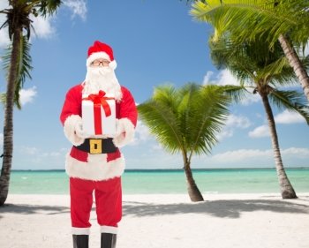 christmas, holidays and people concept - man in costume of santa claus with gift box over tropical beach background