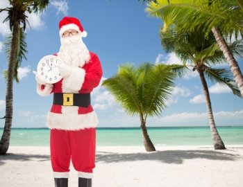 christmas, holidays and people concept - man in costume of santa claus with clock showing twelve pointing finger over tropical beach background