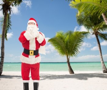 christmas, holidays and people concept - man in costume of santa claus with bag making hush gesture over tropical beach background
