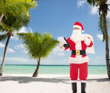christmas, holidays and people concept - man in costume of santa claus with notepad and bag over tropical beach background