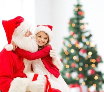holidays, celebration, childhood and people concept - smiling little girl hugging with santa claus over christmas tree lights background