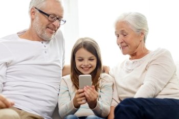 family, generation, technology and people concept - smiling grandfather, granddaughter and grandmother with smartphone sitting on couch at home