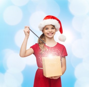 christmas, holidays, happiness and people concept - smiling girl in santa helper hat with gift box and magic wand over blue lights background