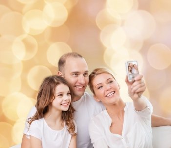 family, holidays, technology and people concept - smiling mother, father and little girl making selfie with camera over beige lights background