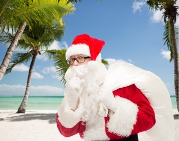 christmas, holidays, travel and people concept - man in costume of santa claus with bag making hush gesture over tropical beach background
