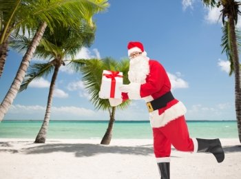 christmas, holidays, travel and people concept - man in costume of santa claus running with gift box over tropical beach background