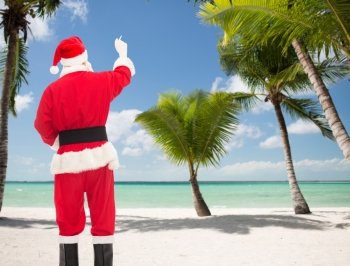 christmas, holidays, travel and people concept - man in costume of santa claus writing something from back over tropical beach background