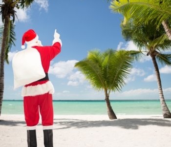 christmas, holidays, travel and people concept - man in costume of santa claus with bag pointing finger from back over tropical beach background