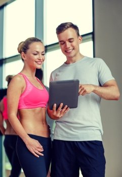 fitness, sport, exercising, technology and diet concept - smiling young woman and personal trainer with tablet pc computer in gym