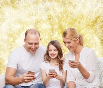 family, holidays, technology and people - smiling mother, father and little girl with smartphones over yellow lights background