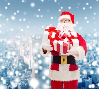 christmas, holidays and people concept - man in costume of santa claus with gift boxes over snowy city background