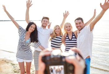 summer, sea, tourism, technology and people concept - group of smiling friends with camera on beach waving hands and photographing