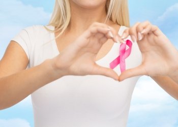 healthcare, people, charity and medicine concept - close up of young woman in blank white t-shirt with pink breast cancer awareness ribbon showing heart shape over blue sky background