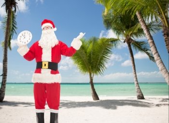 christmas, holidays, travel and people concept - man in costume of santa claus with clock showing twelve over tropical beach background