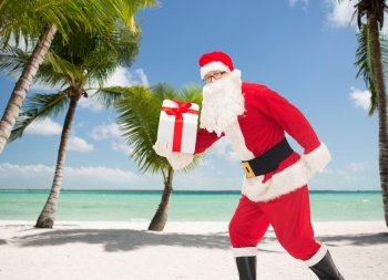 christmas, holidays and people concept - man in costume of santa claus running with gift box over tropical beach background