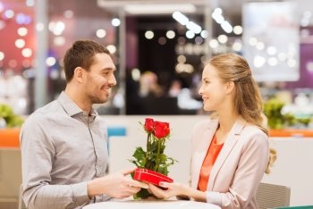 love, romance, valentines day, couple and people concept - happy young man with red flowers giving present to smiling woman at cafe in mall