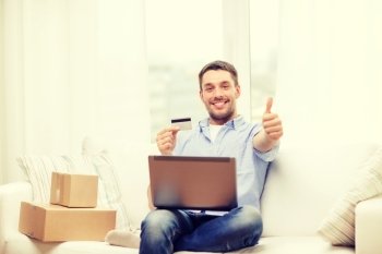 technology, home and lifestyle concept - smiling man with laptop, credit card and cardboard boxes at home showing thumbs up