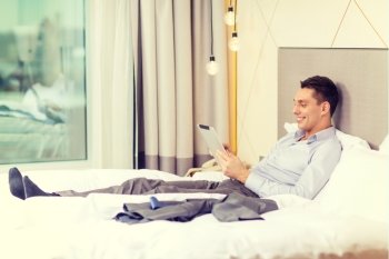 business, technology, internet and hotel concept - happy businessman with tablet pc computer lying in hotel in bed