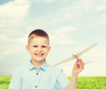 dreams, future, hobby, nature and childhood concept - smiling little boy holding wooden airplane model in his hand over natural background