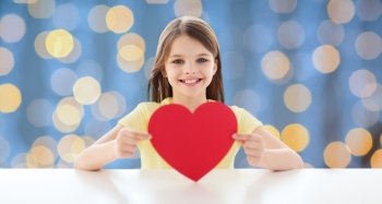 love, charity, holidays, children and people concept - smiling little girl with red heart over blue lights background