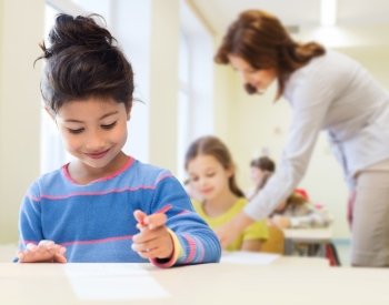 education, elementary school and children concept - happy little student girl with pen and paper writing over classrom and teacher background