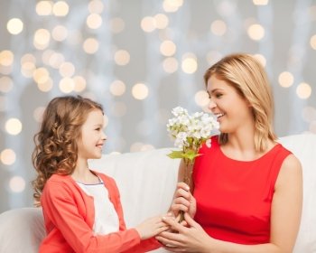 people, holidays, relations and family concept - happy little daughter giving flowers to her mother over holidays lights background