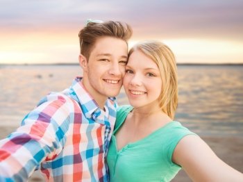 people, love, sea, technology and summer vacation concept - happy couple taking selfie with smartphone or camera over sunset beach background
