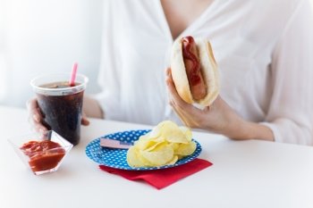 american independence day, celebration, patriotism and holidays concept - close up of woman hands holding hot dog and coca cola in plastic cup with potato chips and ketchup on 4th july at home party
