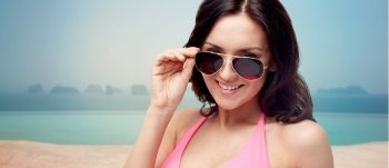 people, fashion, travel, tourism and summer concept - happy young woman in sunglasses and pink swimsuit looking at you over infinity pool at sea side background