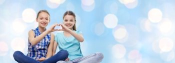people, children, friends and friendship concept - happy little girls sitting and showing heart shape hand sign over blue holidays lights background
