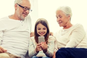 family, generation, technology and people concept - smiling grandfather, granddaughter and grandmother with smartphone sitting on couch at home