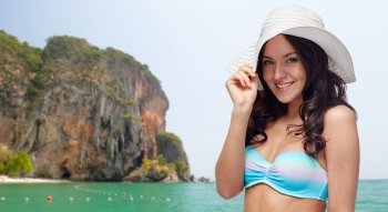 people, fashion, summer and beach concept - happy young woman in bikini swimsuit and sun hat over sea and rock background