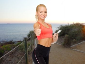 sport, fitness, technology, gesture and people concept - smiling sporty woman smartphone showing thumbs up over beach sunset background