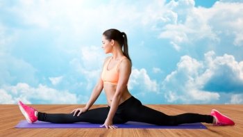fitness, sport, exercising, stretching and people concept - smiling woman doing splits on mat over wooden floor and sky with white clouds background