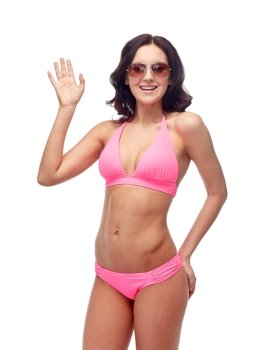 people, fashion, swimwear, summer beach and gesture concept - happy young woman in sunglasses and pink swimsuit waving hand