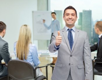business, people, gesture and success concept - happy smiling businessman with team over office room background showing thumbs up