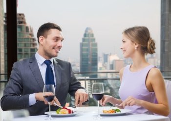 people, eating, celebration, romantic and holidays concept - smiling couple with red wine and food talking at restaurant over city background