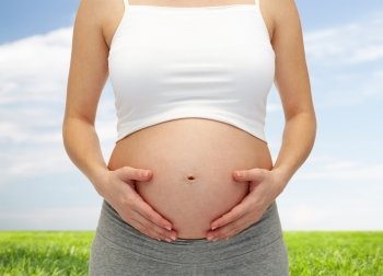 pregnancy, motherhood, people and expectation concept - close up of pregnant woman touching her bare tummy over blue sky and grass background