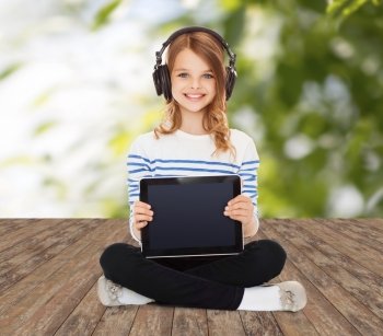music, technology, people and childhood concept - happy girl with headphones showing tablet pc computer blank screen over greed background