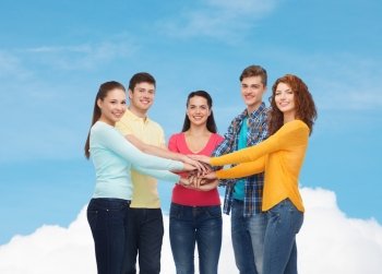 friendship, dream, teamwork, gesture and people concept - group of smiling teenagers with hands on top of each other over blue sky with white cloud background