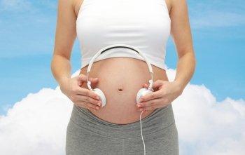 pregnancy, people, music, technology and expectation concept - close up of happy pregnant woman applying headphones to bare tummy over blue sky and cloud background