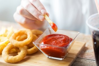 fast food, people and unhealthy eating concept - close up of hand with deep-fried squid rings, dipping french fries into ketchup bowl on wooden table