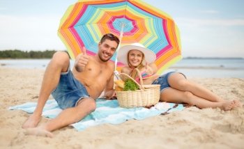 summer, holidays, vacation and happy people concept - happy couple with picnic basket and parasol showing thumbs up on beach