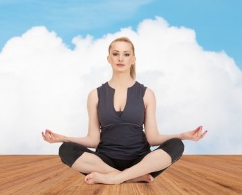 people, health, wellness and meditation concept - happy young woman meditating in yoga lotus pose on wooden floor over white cloud and blue sky background