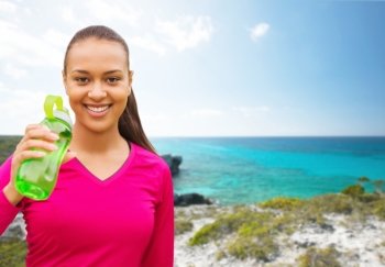fitness, people and sport concept - smiling african american woman drinking from bottle over beach background