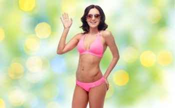 people, fashion, swimwear, summer beach and gesture concept - happy young woman in sunglasses and pink swimsuit waving hand over green holidays lights background