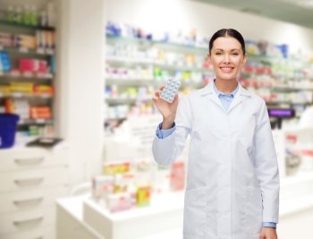 medicine, pharmacy, people, health care and pharmacology concept - happy young woman pharmacist with pills over drugstore background