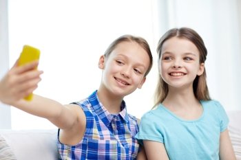 people, children, technology, friends and friendship concept - happy little girls sitting on sofa and taking selfie with smartphone at home